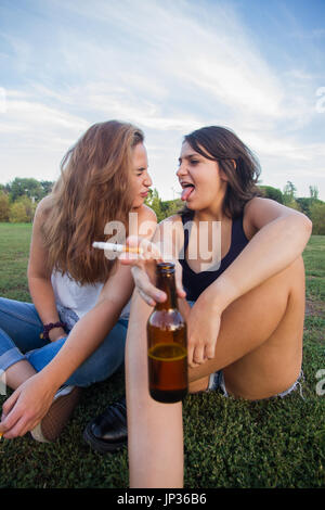 Two girls, friends, smoking cigarettes and drinking beer bottles in the park on a cloudy day. They are of celebration and are happy. Stock Photo