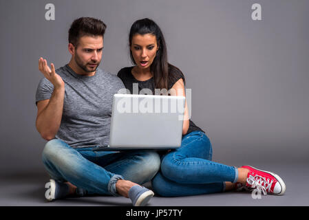 A beautiful young couple sitting on the ground and looking at a laptop and feeling surprised Stock Photo