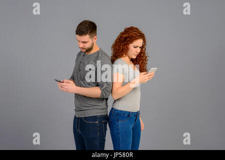 A beautiful young couple standing back to back while holding their smartphones and feeling surprised Stock Photo