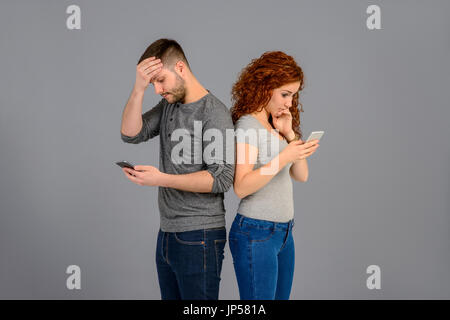 A beautiful young couple standing back to back while holding their smartphones and feeling surprised Stock Photo