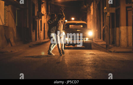 A man and a woman dancing together in front of a classic 1950s car in a street at night. Stock Photo