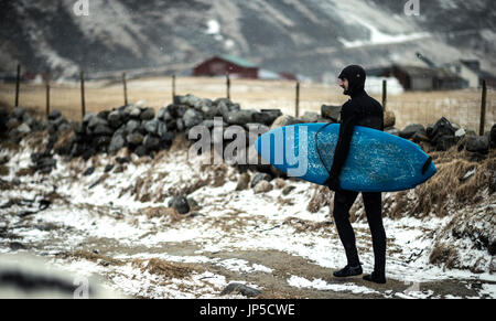 A surfer wearing a wetsuit and carrying a surfboard walking along a snowy beach. Stock Photo