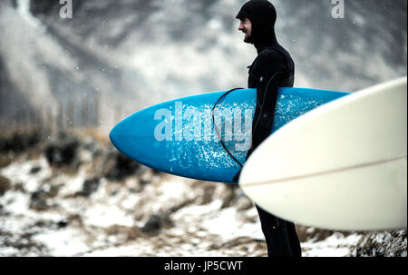 A surfer wearing a wetsuit and carrying surfboards standing on a snowy beach with mountains behind. Stock Photo