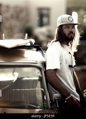 Bearded young man wearing baseball cap leaning against a parked car. Stock Photo