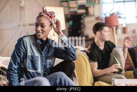 Young woman wearing headscarf and young man holding beer bottle sitting indoors on a sofa, smiling. Stock Photo