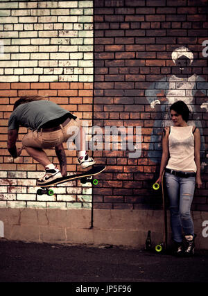 A young man and woman skateboarding in front of a brick wall. Stock Photo