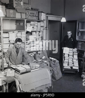 1950s, picture shows a coated employee with tie packing books up ready for despatch in a store room at the famous bookshop of W & G Foyle Ltd, Charing Cross Rd, London, while another suited employee brings more books in on a trolley. Stock Photo