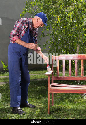 Senior man in overalls painting old bench in garden after sandblasting. Repairing old furniture Stock Photo
