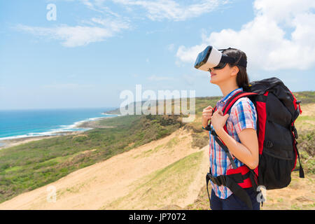 smiling female backpacker wearing virtual reality device experience 3D video with coastal scenery standing on hiking mountain with personal baggage. Stock Photo