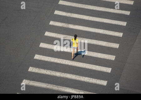 Aerial view of woman in yellow top walking with shadow cast across lines of crosswalk of a wide street Stock Photo