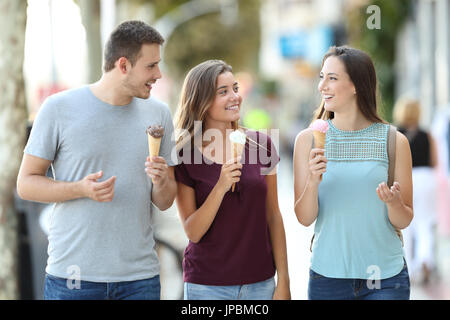 Front view of three happy friends talking and eating ice creams walking in the street Stock Photo