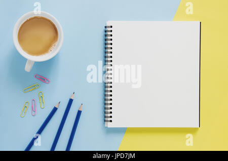 Office workplace minimal concept. Blank notebook, cup of coffee, pencil, paper clip on yellow and blue background. Top view with copy space, flat lay. Stock Photo