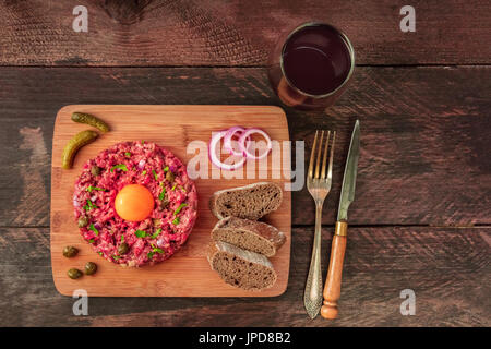 A photo of a steak tartare with a raw egg yolk, gherkins, capers, rye bread, purple onions, a glass of red wine, a fork, and a knife, shot from above  Stock Photo