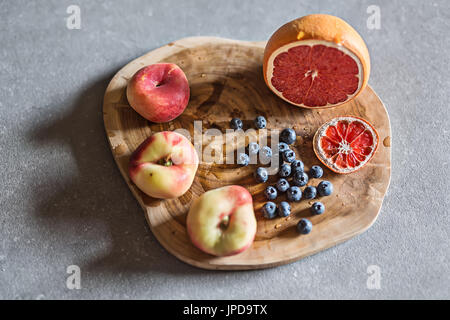 Colorful fruit on the textured wooden board on the gray surface. There is a heap of blueberries, three peaches and a sliced grapefruit. Closeup top vi Stock Photo
