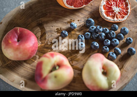 Multicolored fruit on the textured wooden board on the gray surface. There is a heap of blueberries, three peaches and a sliced grapefruit. Closeup. H Stock Photo
