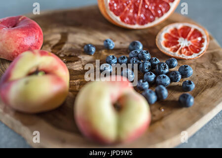 Fresh and juicy fruit on the textured wooden board on the gray surface. There is a heap of blueberries, three peaches and a sliced grapefruit. Closeup Stock Photo