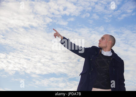 Bald young man with brown eyes dressed in modern and stylish clothes on blue sky background with clouds. Stock Photo