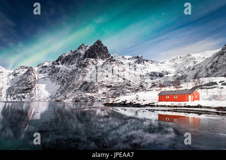 Lofoten, Svoelver, Aurora Borealis over a frozen lake and red rorbu, Norway.