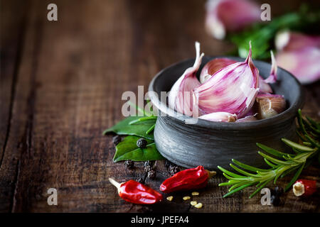 Cloves of purple garlic in a black bowl with rosemary, bay leaf, black pepper and chili pepper on dark rustic wooden background. With plenty of copy s Stock Photo