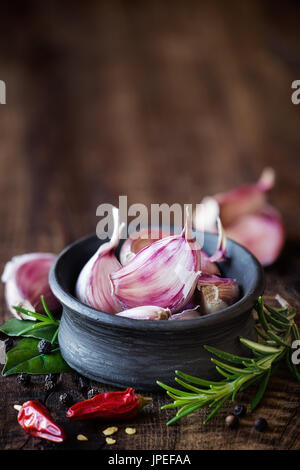 Cloves of purple carlic in a black bowl with rosemary, bay leaf, black pepper and chili pepper on dark rustic wooden background. With plenty copy spac Stock Photo