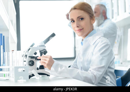 caucasian chemist in white coat working with microscope and reagents while looking at camera  in chemical laboratory   Stock Photo