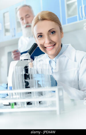 caucasian smiling scientist in white coat working with microscope and reagents while looking at camera with colleague behind in lab Stock Photo