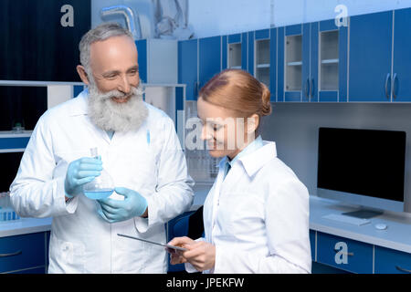 smiling chemists in lab coats holding flask and digital tablet at laboratory Stock Photo