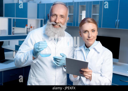 smiling chemists in lab coats looking at camera and holding flask and digital tablet Stock Photo
