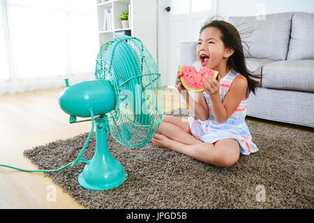 beautiful youth girl holding watermelon sitting in front of electric fan and blowing cool fan for eliminating summer hot. Stock Photo