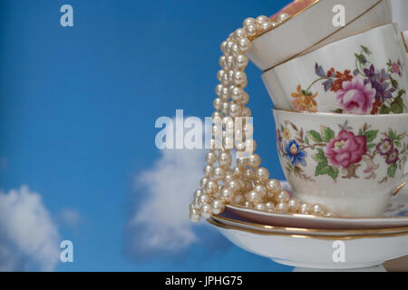 still life arrangement with a pile of vintage tea cups and saucers and pearl necklace against a blue sky