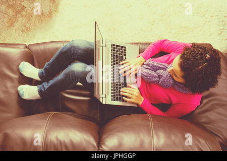 Short curly haired girl is using laptop at home. She is comfortable on a brown leather couch and wearing fall clothes with a scarf. vintage filter. To Stock Photo