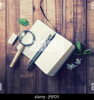 Blank notebook, magnifier, pencil, pen, eraser, flowers and leaves on vintage wood background. Stationery elements. Responsive design mockup Stock Photo