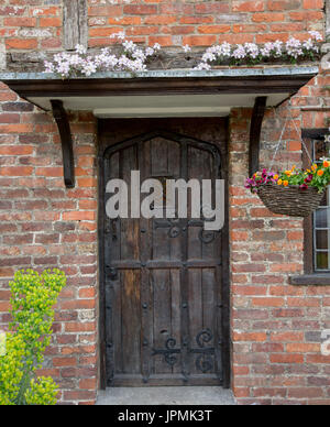 Dark brown wooden door, of historic cottage, with decorative hinges, in red brick wall draped with clematis & hanging basket of colourful flowers Stock Photo