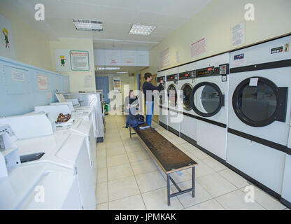 Two men in laundromat, one reading magazine and waiting for washing to be completed, one standing and operating washing machine, Stock Photo