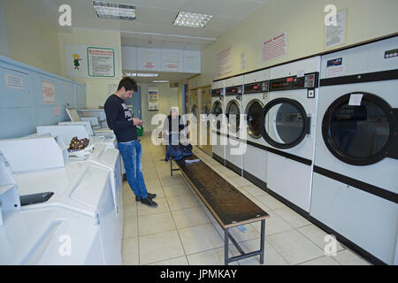 Two men, one reading magazine and one using phone, waiting in laundromat for washing to be completed Stock Photo