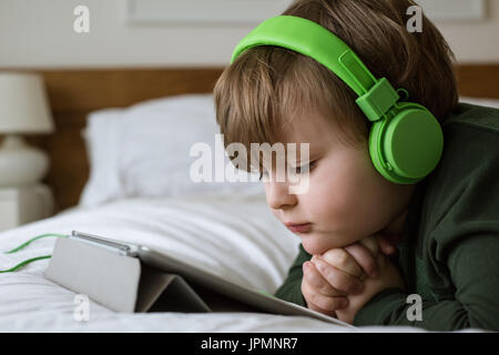 Close up of cute boy wearing headphones while using digital tablet on bed at home Stock Photo