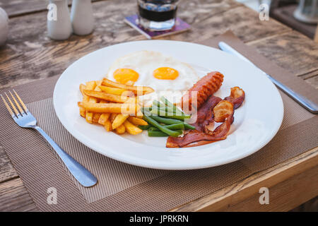 English breakfast with fried eggs, bacon, sausages, green beans and potato french fries on wooden table Stock Photo