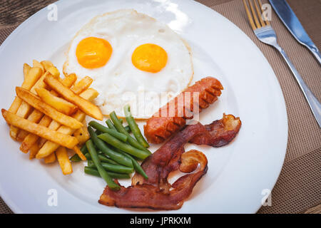 English breakfast with fried eggs, bacon, sausages, green beans and potato french fries on wooden table Stock Photo