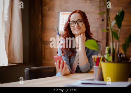 Young woman working from home. Red haired freelancer woman sitting at working place, smiling, looking in camera. Toned image. Stock Photo