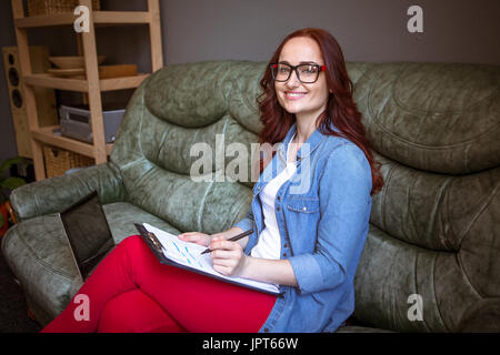Red haired businesswoman working at home. Female sitting on sofa correcting documents. Stock Photo
