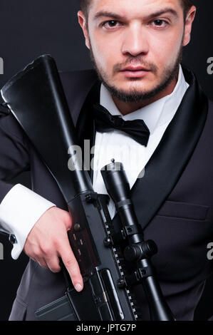 Portrait of young gangster holding armed rifle on dark background as danger and violence concept Stock Photo