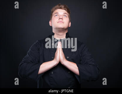 Talented young chef with palms clasped praying and looking up isolated on balck background Stock Photo