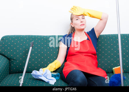 Exhausted young cleaner with headache  sitting on couch at home after cleaning the apartment Stock Photo