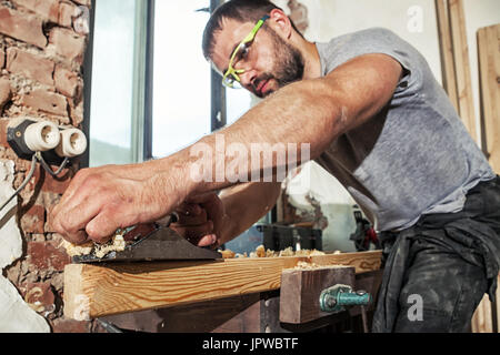 A dark-haired man with a beard and protective green glasses treating a wooden bar with a black jack plane, work with wooden Stock Photo