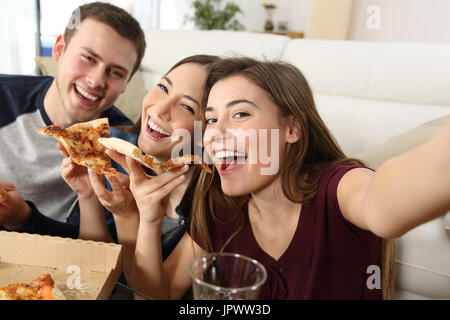 Three happy friends taking selfies and eating pizza sitting on a sofa at home Stock Photo