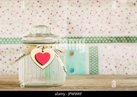 Empty glass jar woth handmade wooden heart decoration and ribbon on a wooden plank with colorful pastel background. Stock Photo