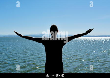 the silhouette of a young caucasian man seen from behind with his arms in the air in front of the ocean, feeling free Stock Photo