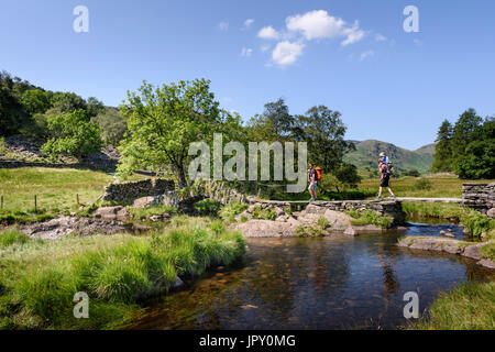 Two fathers carrying children in baby carriers over Slater's Bridge & River Brathay in Little Langdale in the English Lake District, Cumbria Stock Photo