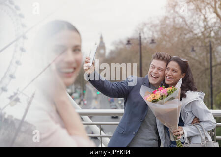 Enthusiastic couple with flower bouquet taking selfie with camera phone on urban bridge, London, UK