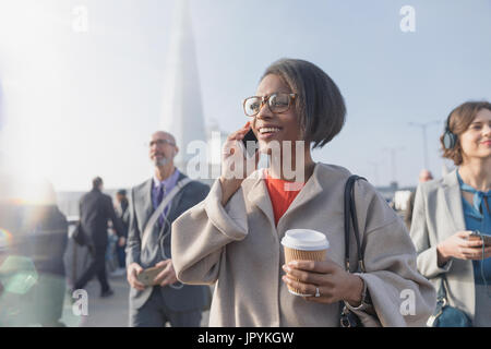 Smiling businesswoman drinking coffee and talking on cell phone on sunny busy urban pedestrian bridge Stock Photo